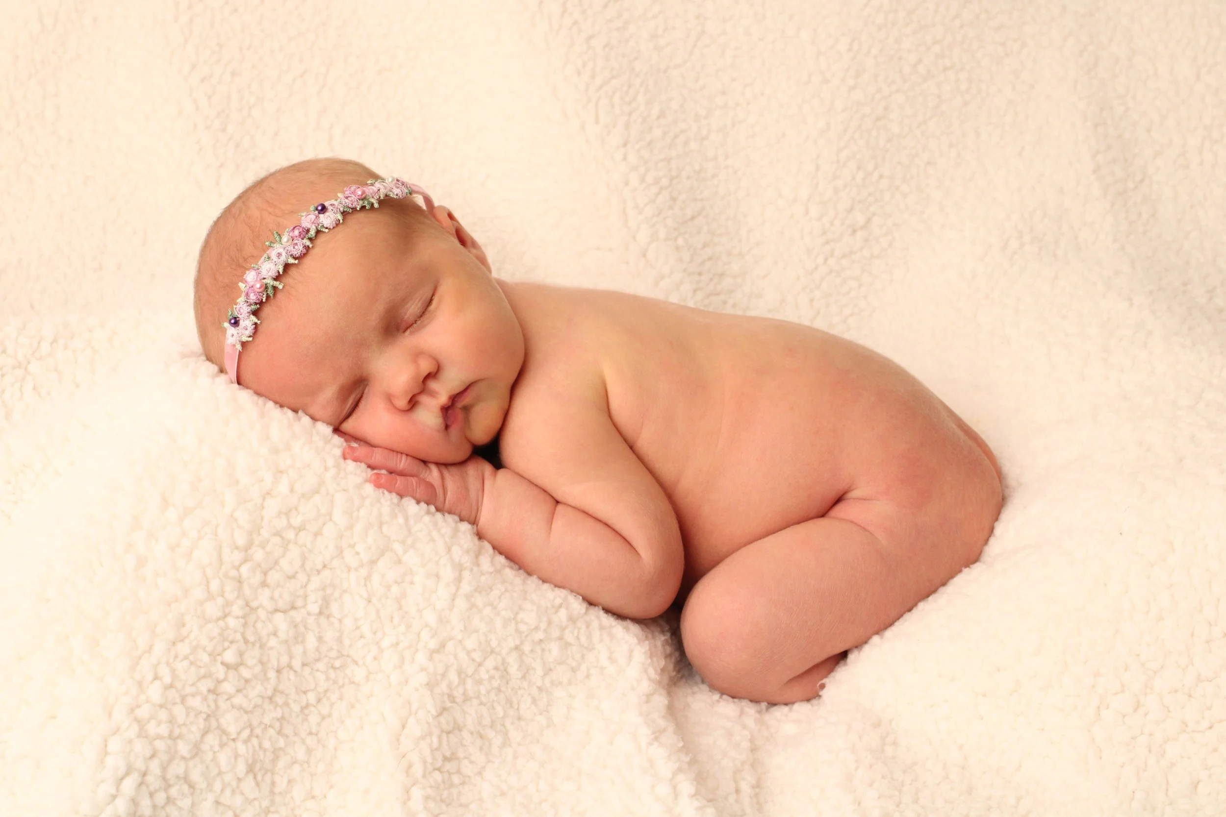 Newborn baby girl wearing a pink floral headband, sleeping on a cream blanket during a newborn photography session