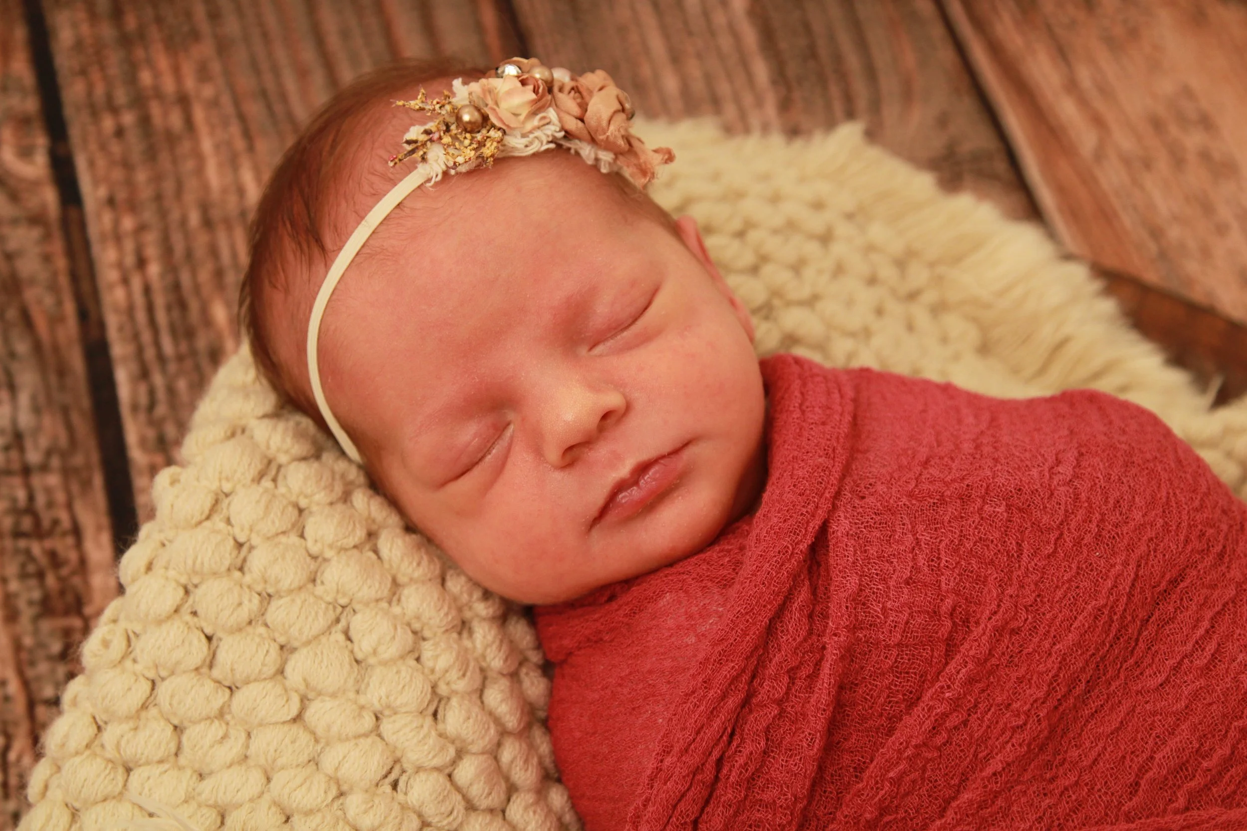 Newborn baby girl wrapped in a red knit wrap with a floral headband, photographed against a rustic wood backdrop